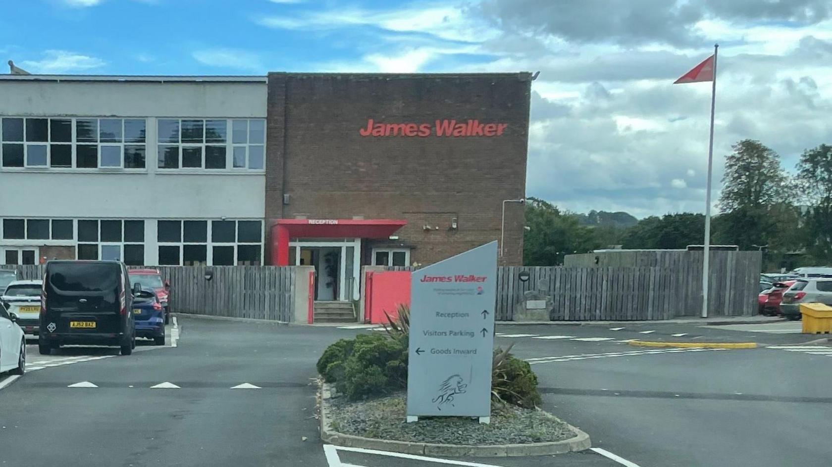 Photo from car park of James Walker building. Large brick building has "James Walker" written in red on it. There is a sign in the car park directing cars and a flagpole in the right of the picture.
