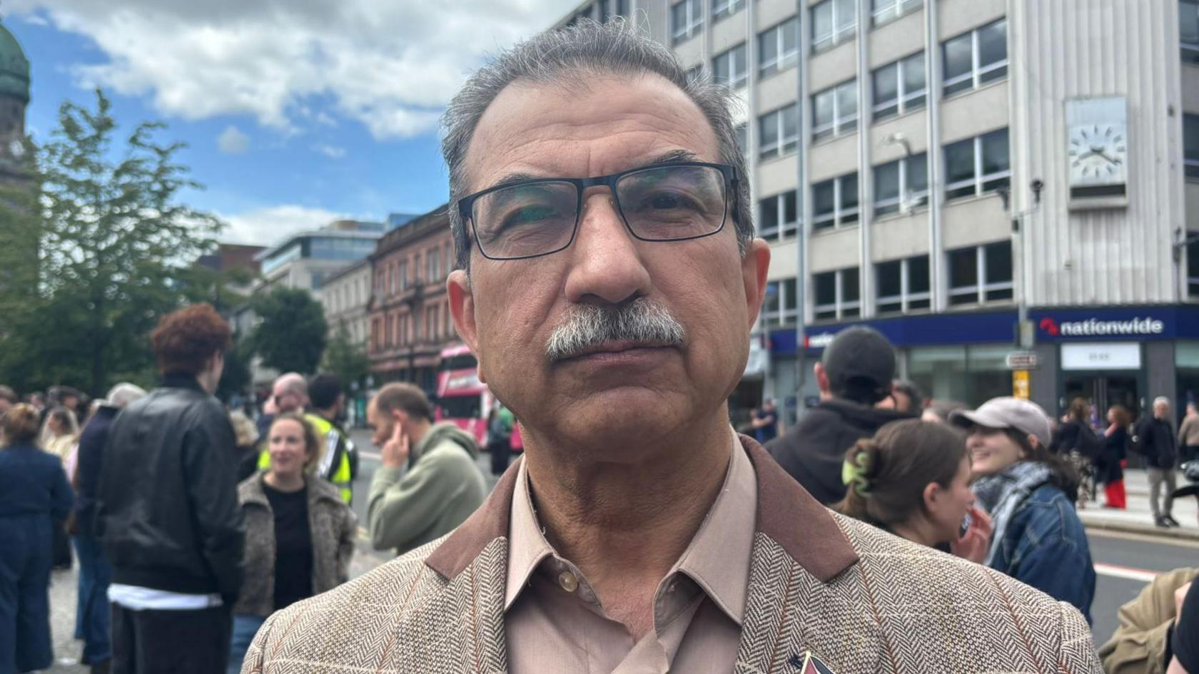 Raied is looking directly at the camera. He is wearing a tweed blazer and a nude coloured shirt. He has grey hair and square-framed glasses. 
