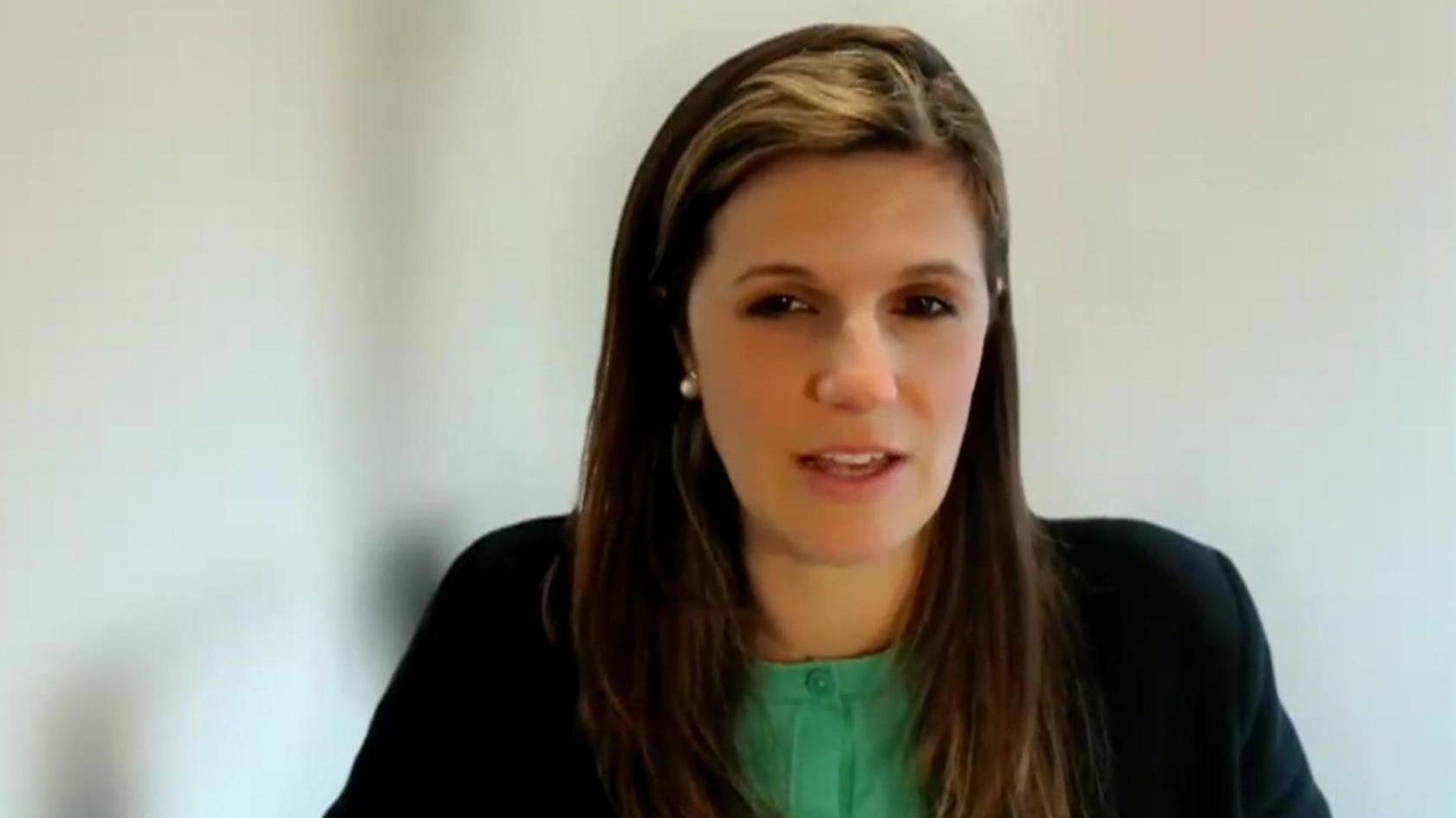 Clare Fernyhough sitting facing the camera on a video call, in front of a blurred background. She has long dark hair and is wearing a green top with black jacket