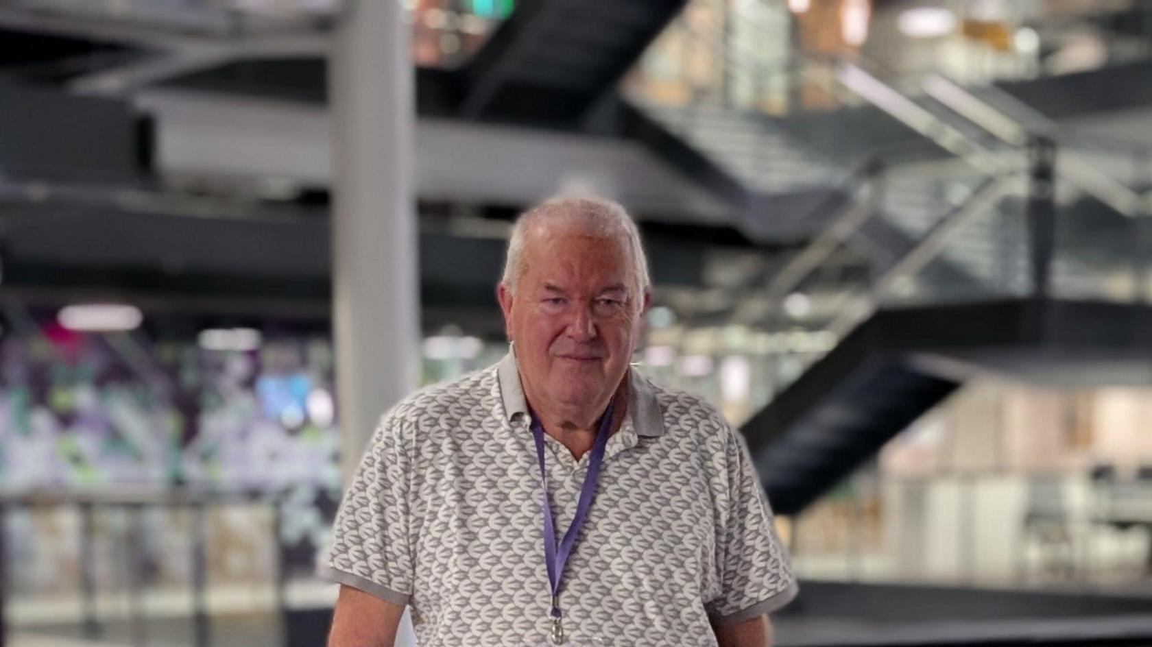 Steve Mitchell, has silver short hair, and stands centre infront of the glass rail on the first floor inside BBC central square building in Cardiff. He wears a white and grey polo shirt and has a purple visitor lanyard around his neck. 