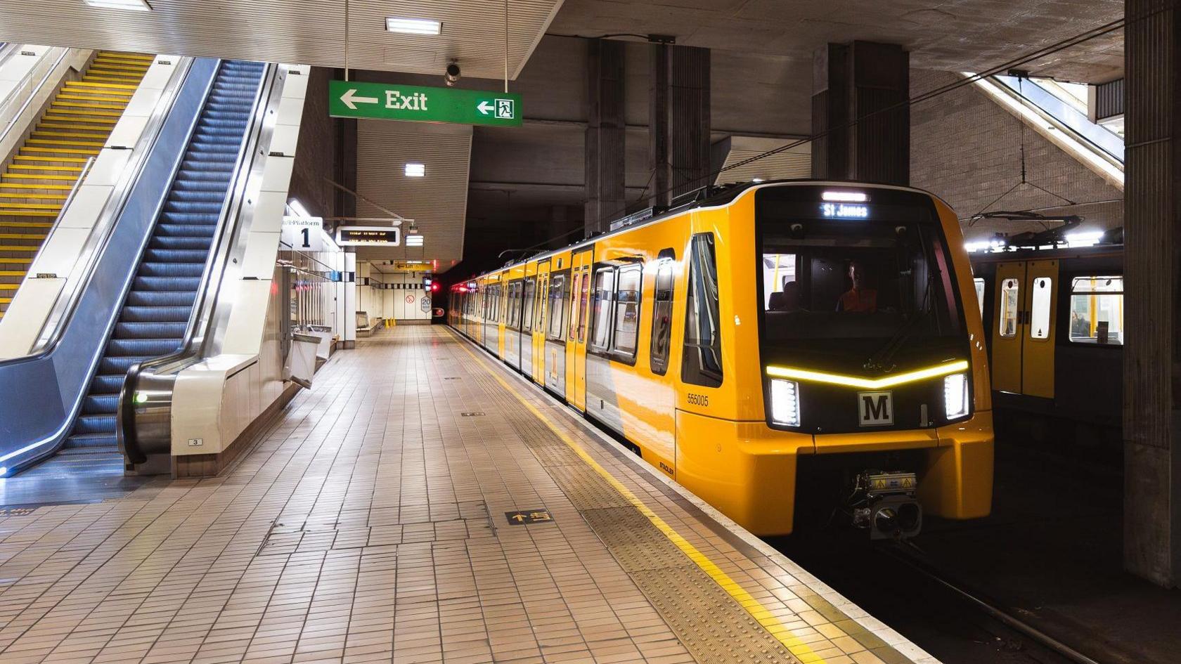 A new Metro train, which is shiny yellow and grey, at St James' station. The platform is beige tiled and there are escalators to the side.