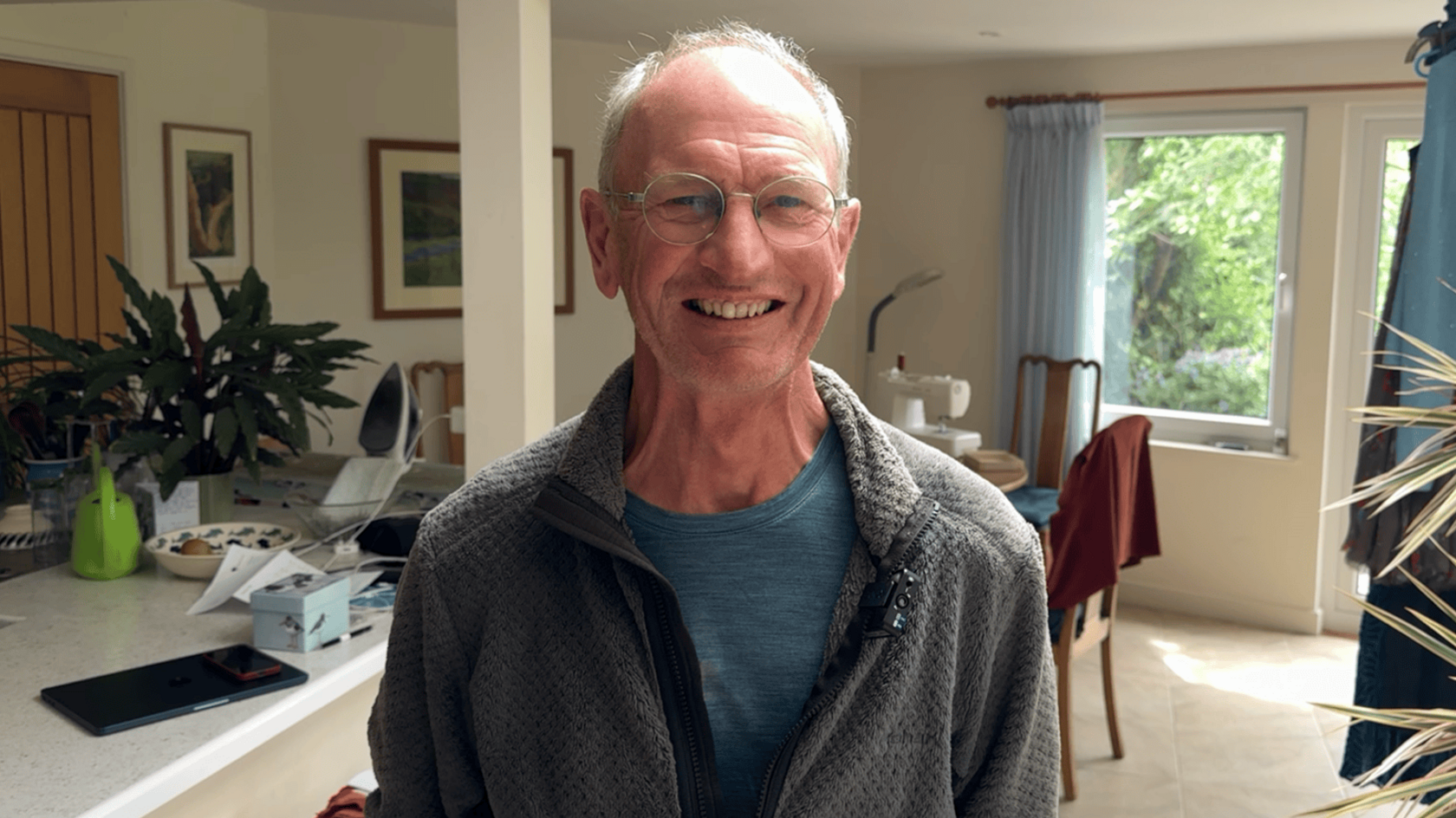 Andy Cook is pictured. He is a white man in his 60s and he is smiling at the camera. He has receding grey hair and is wearing a blue T-shirt and grey fleece. He is standing in the kitchen of his home.