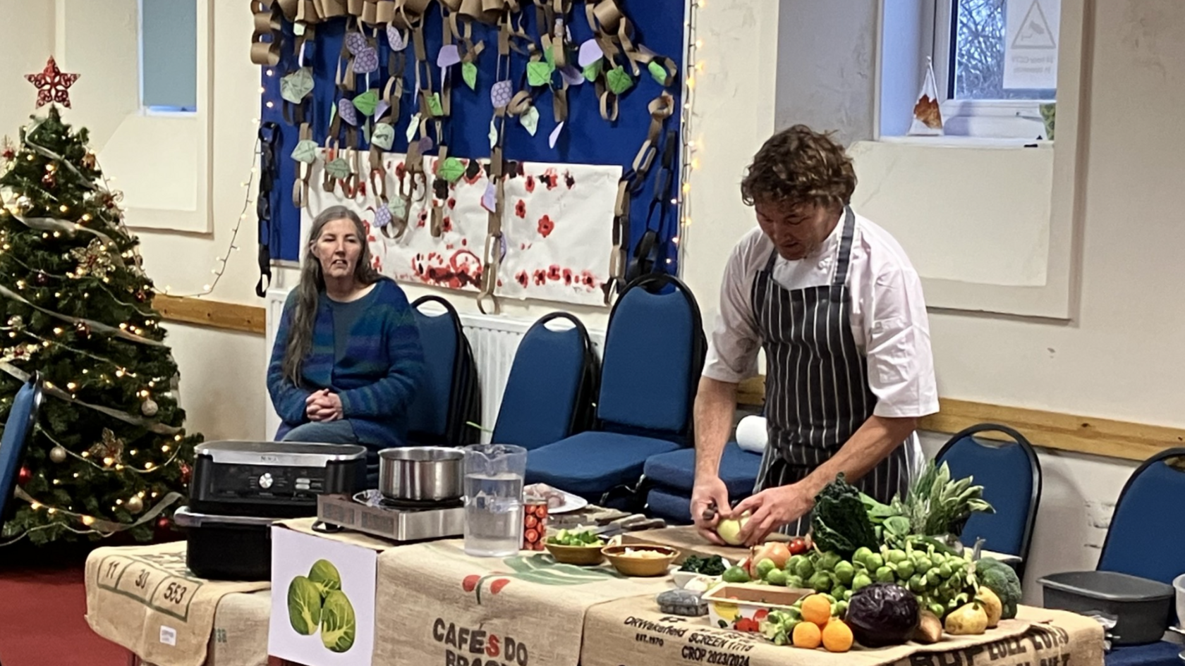 A chef, in a white shirt and a black and white striped apron, cutting an onion. There are different vegetables on the table. There is a Christmas tree to the left and a woman sat on a blue chair waiting blue. 