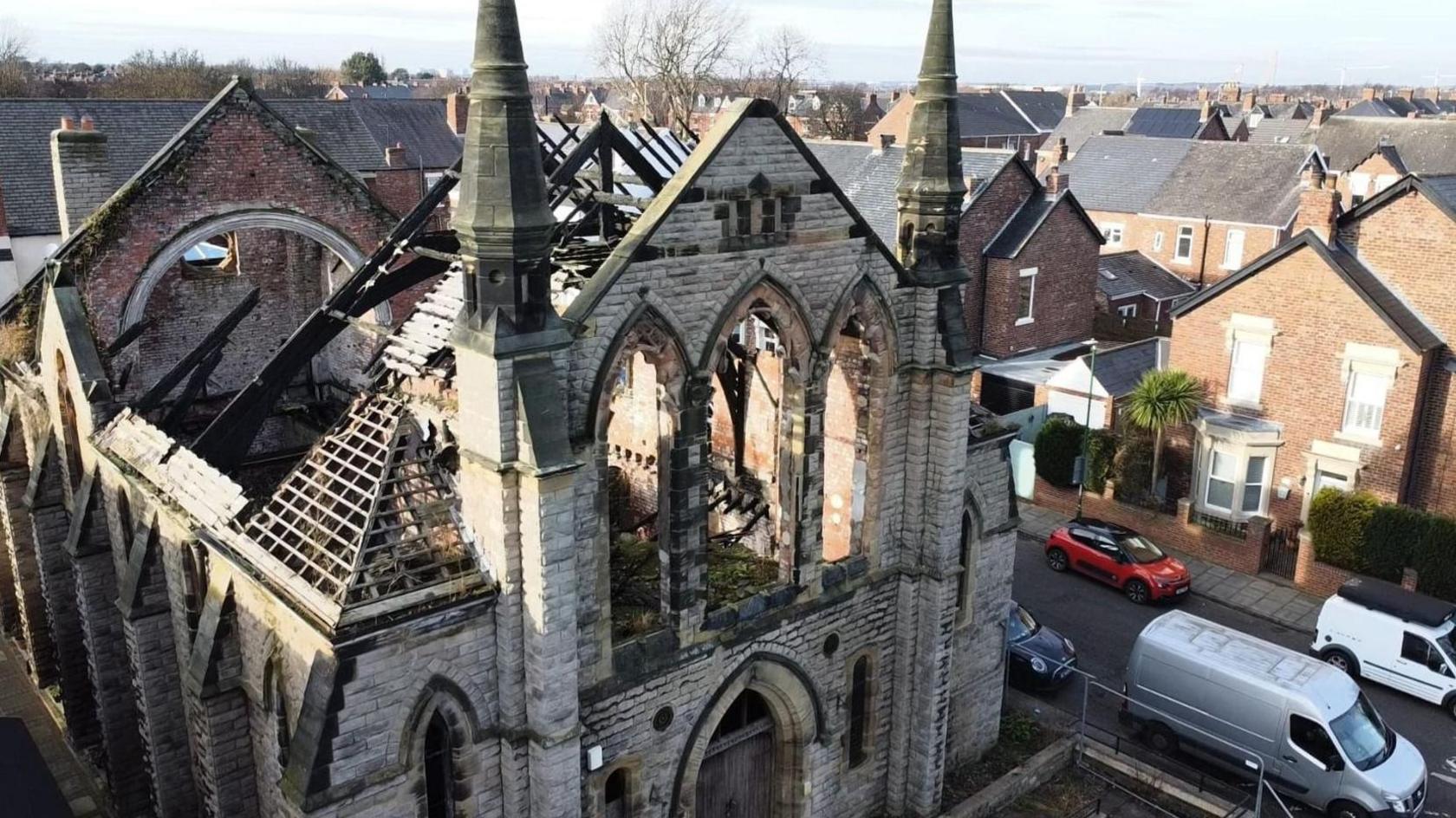 An old church building that does not have a roof. It is the shell of a building. There are houses nearby.