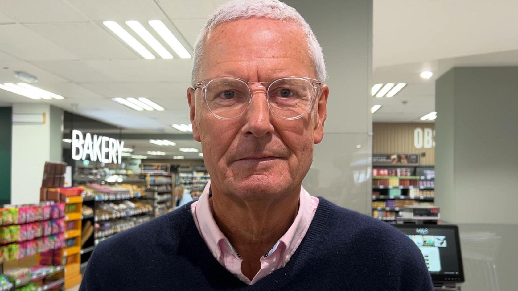Sandpiper CI executive chairman Tony O'Neill looks at the camera with a neutral look on his face. He is wearing a navy blue jumper over a pink shirt. He has clear framed glasses and short grey hair. He is stood inside a Marks and Spencer store with several aisles full of stock and a till behind him. A sign saying "bakery" is lit up in the background.