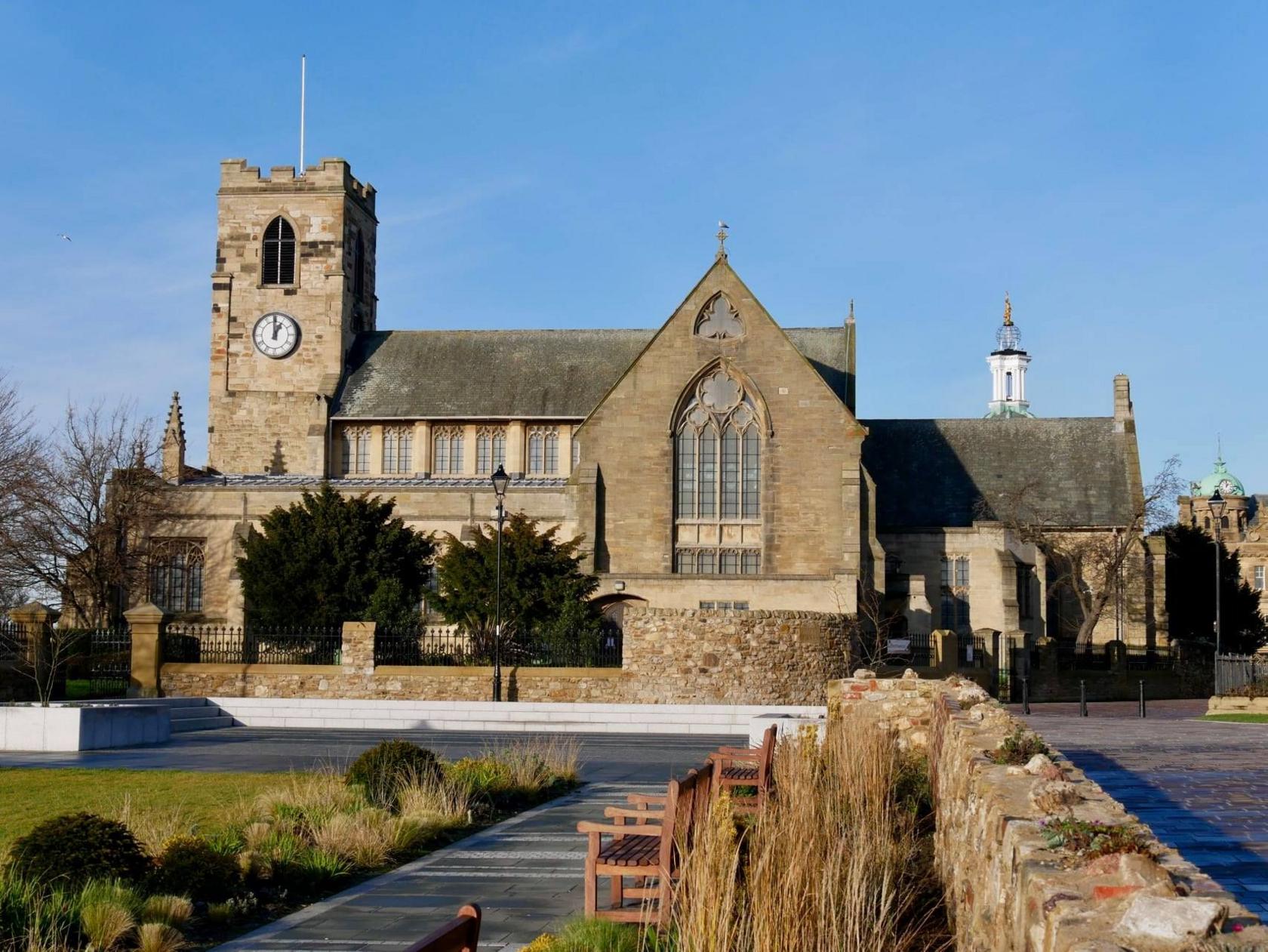 Sunderland Minster: a greystone church with large, imposing windows and a clock tower. A courtyard with benches and greenery lie in front.