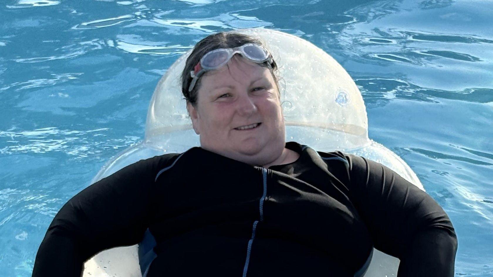 Gail is lying on an inflatable in a swimming pool. She is wearing a black swimming costume and has googles sitting on her head. 