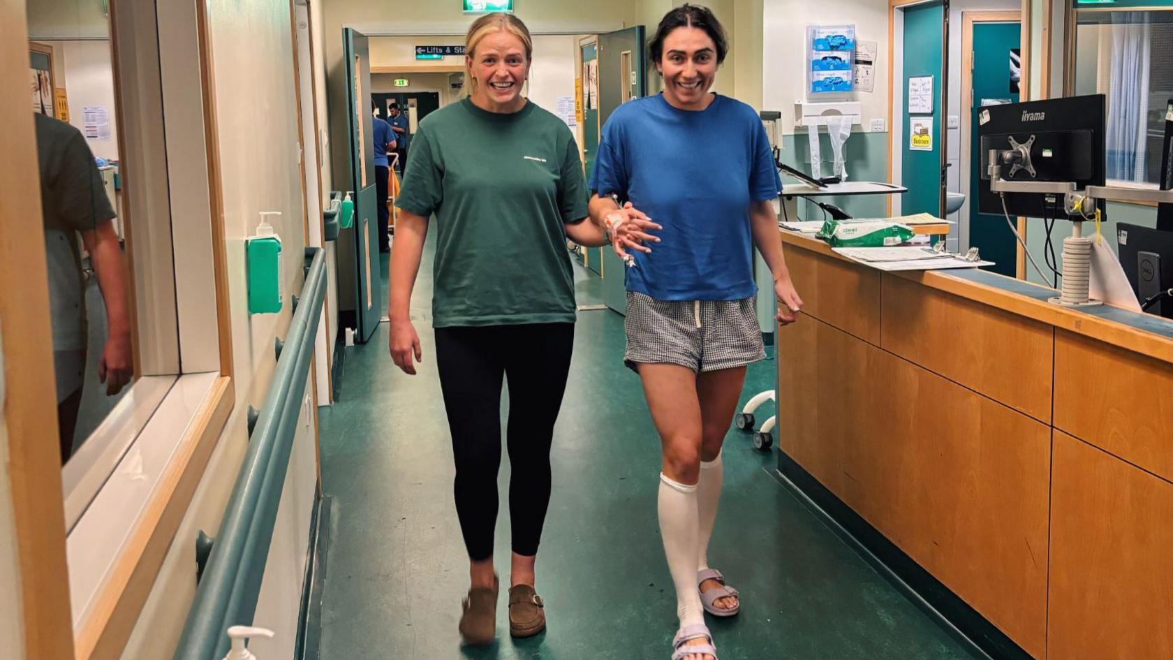 Emma Wassell in hospital but going for a walk, dressed in a blue shirt and grey shorts with a friend dressed in a green top