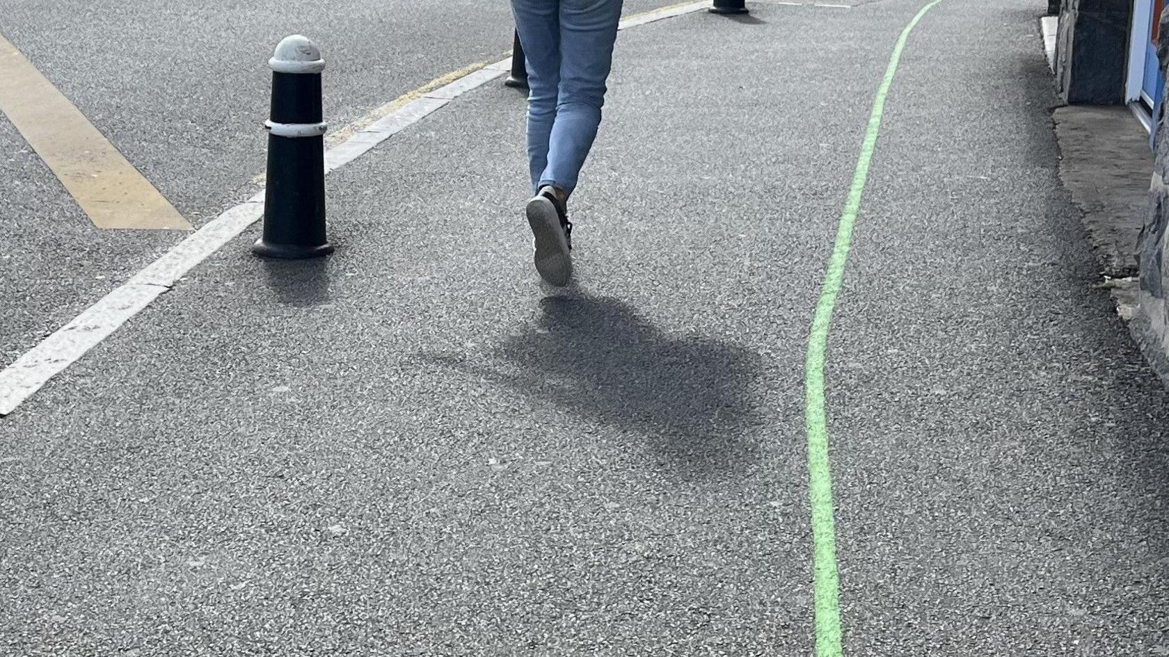 Green line running along a pavement. A pedestrian walks next to the line away from the camera - only their legs in jeans and feet in trainers can be seen - and their shadow can be seen behind them. A bollard on the left of the picture is at the edge of the pavement next to a yellow line.