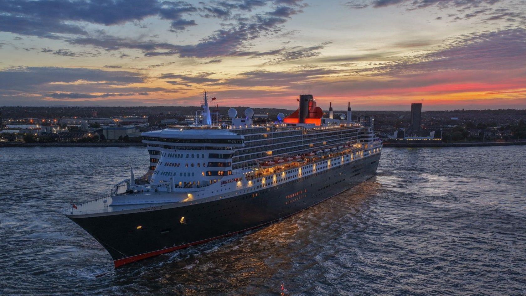 The Queen Mary 2 ocean liner on the River Mersey at night. The Wirral peninsula is in the background and the sky is purple and yellow as the sun sets.