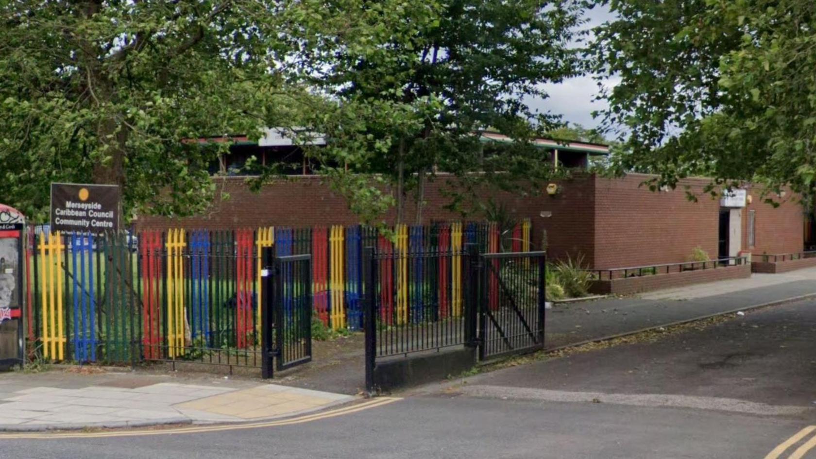 A single storey brown brick building with a metal fence painted red, yellow, blue, green and black. A brown sign with white writing reads Merseyside Caribbean Council Community Centre