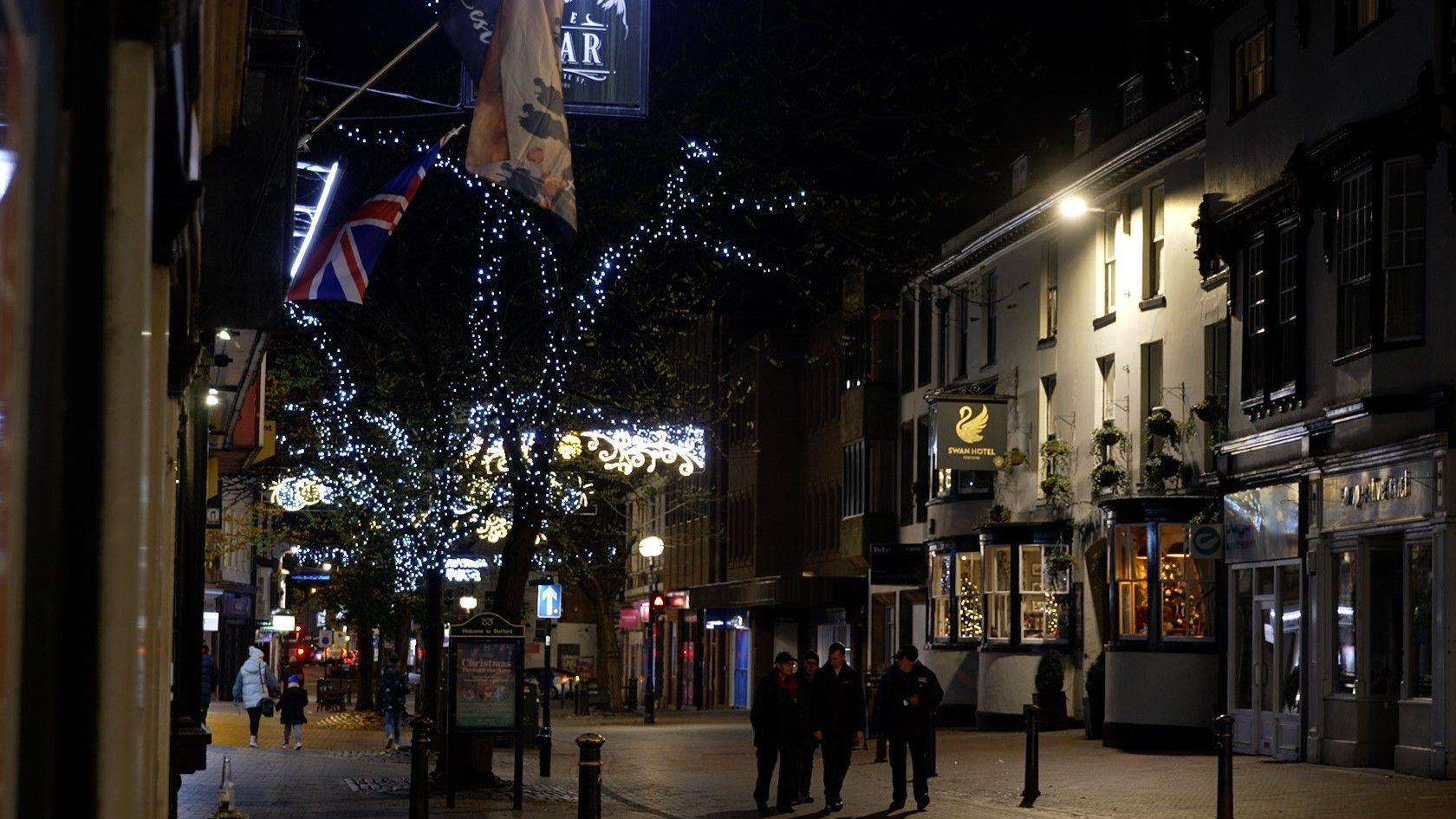 Stafford High Street at night, with trees lit up in Christmas lights, shop windows and bar entrances along the pedestrian area. There are people walking around the town.