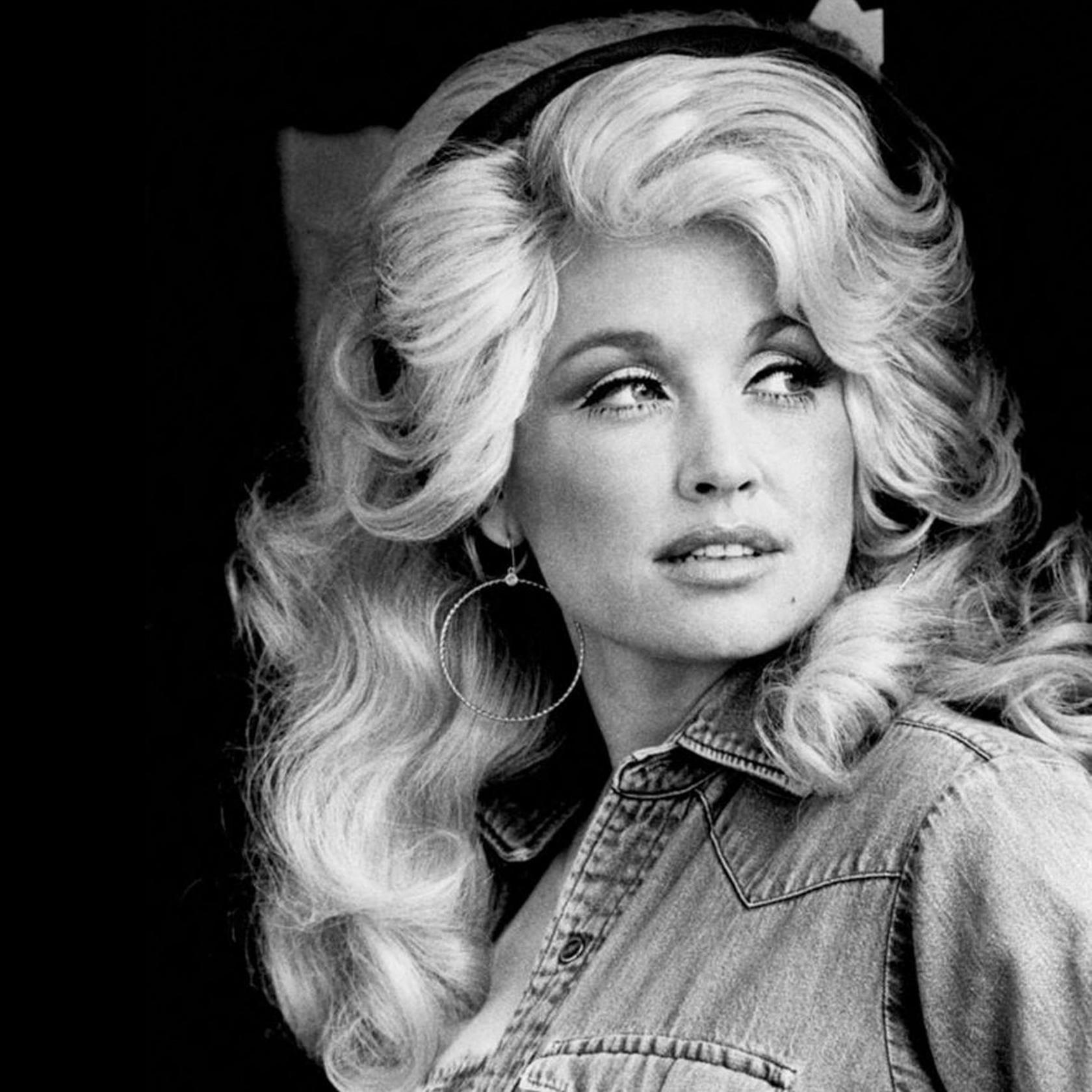 Dolly Parton, with big blond curls, hoop earrings, and a denim shir in a black and white portrait from the 1970s