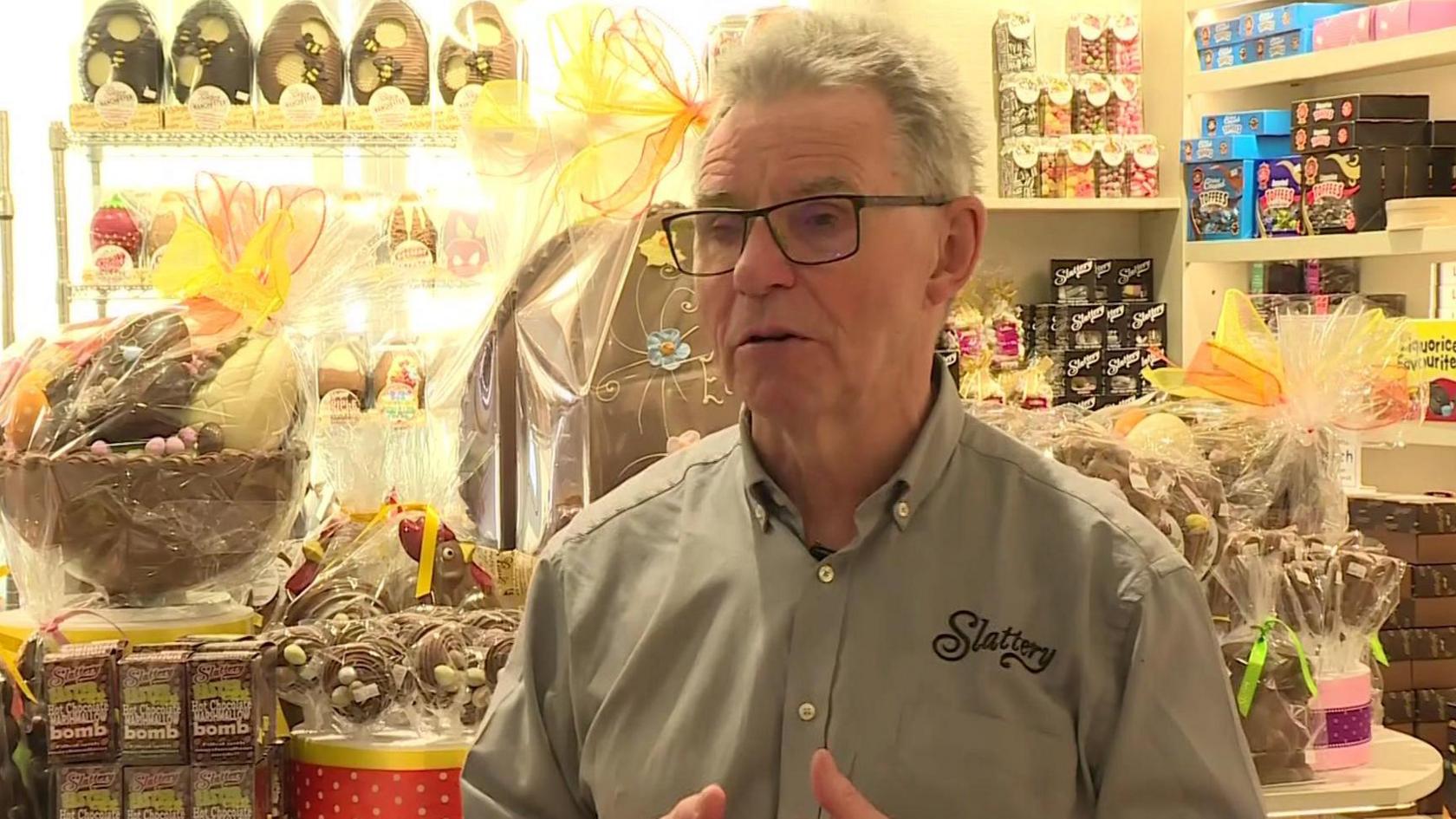 John Slattery, standing in front of lots of chocolate. He has grey hair, is wearing glasses and a light buttoned shirt with Slaterry printed on.