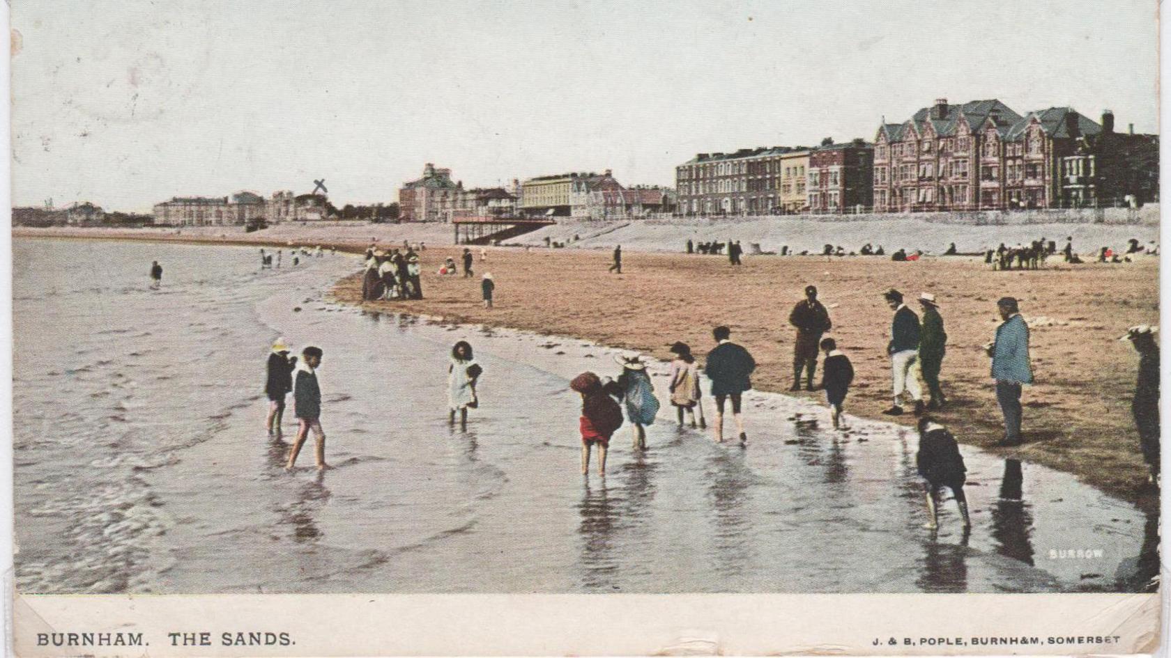 An old postcard showing Burnham's beach. People are walking through the water. In the background are houses along the promenade.