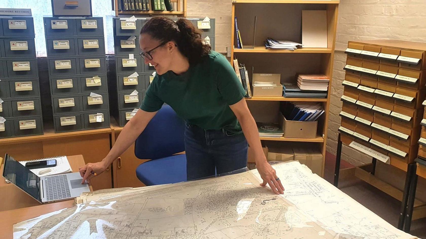 Katie Owen, a local studies adviser working with the archive, stands over an OS map spread across a table while moving her finger across a trackpad on a laptop. 