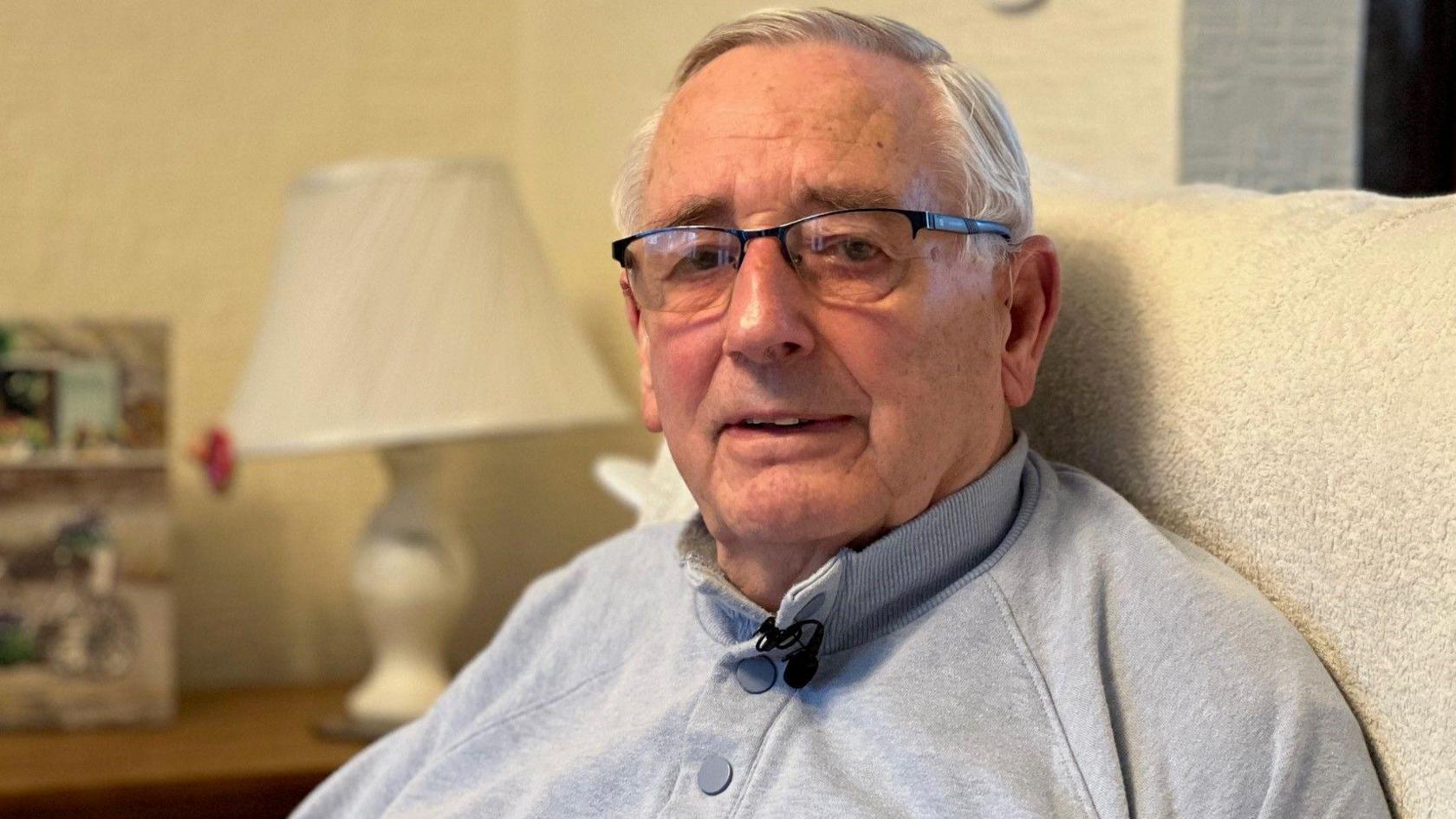 Colin Gray wearing a grey jumper sits in a beige chair