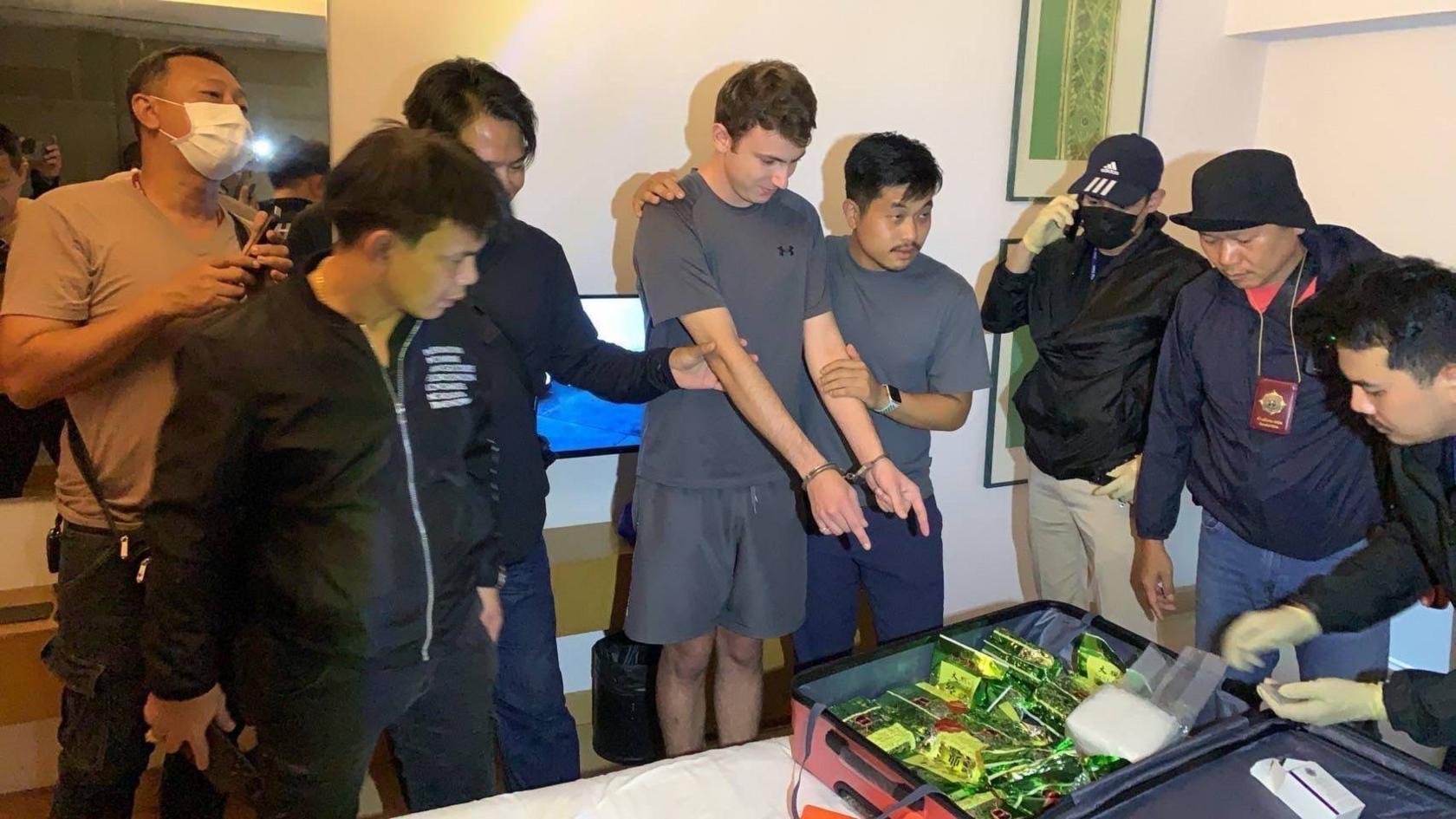A man in a grey shirt and grey shorts. He is looking down and pointing at a suitcase on a bed containing packages in green foil. He is handcuffed. He is surrounded by seven other men - they appear to be Thai police officers in plain clothes. 