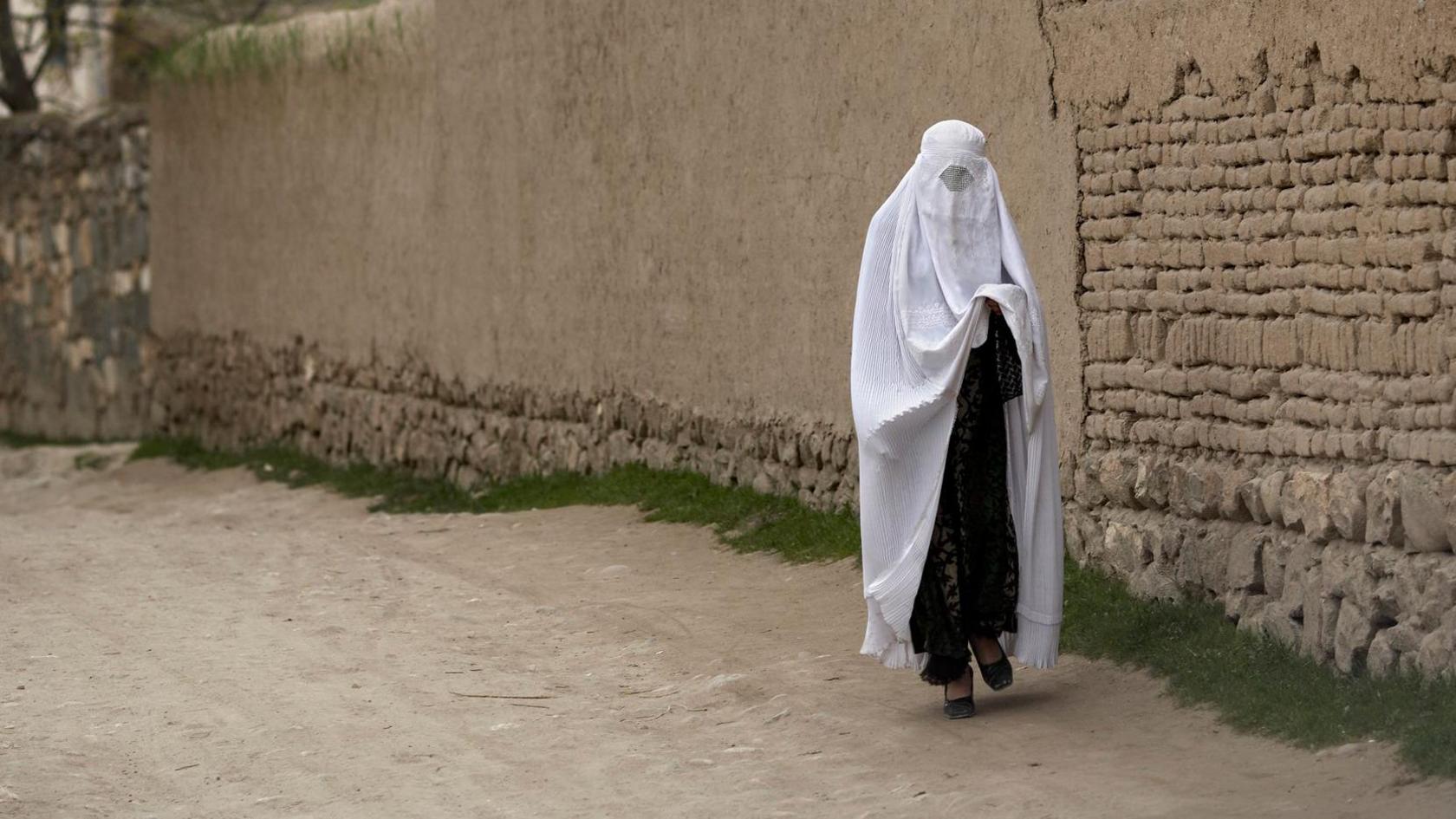 A woman wearing a white burka - a full veil - walking along a road in Afghanistan