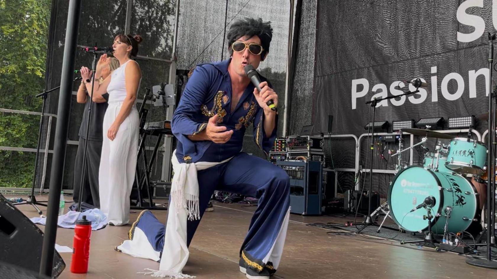A man dressed as Elvis Presley with big glasses and hair singing into the camera, with the majority of the stage behind him. Also on the stage are two women backing singers and drum kit.