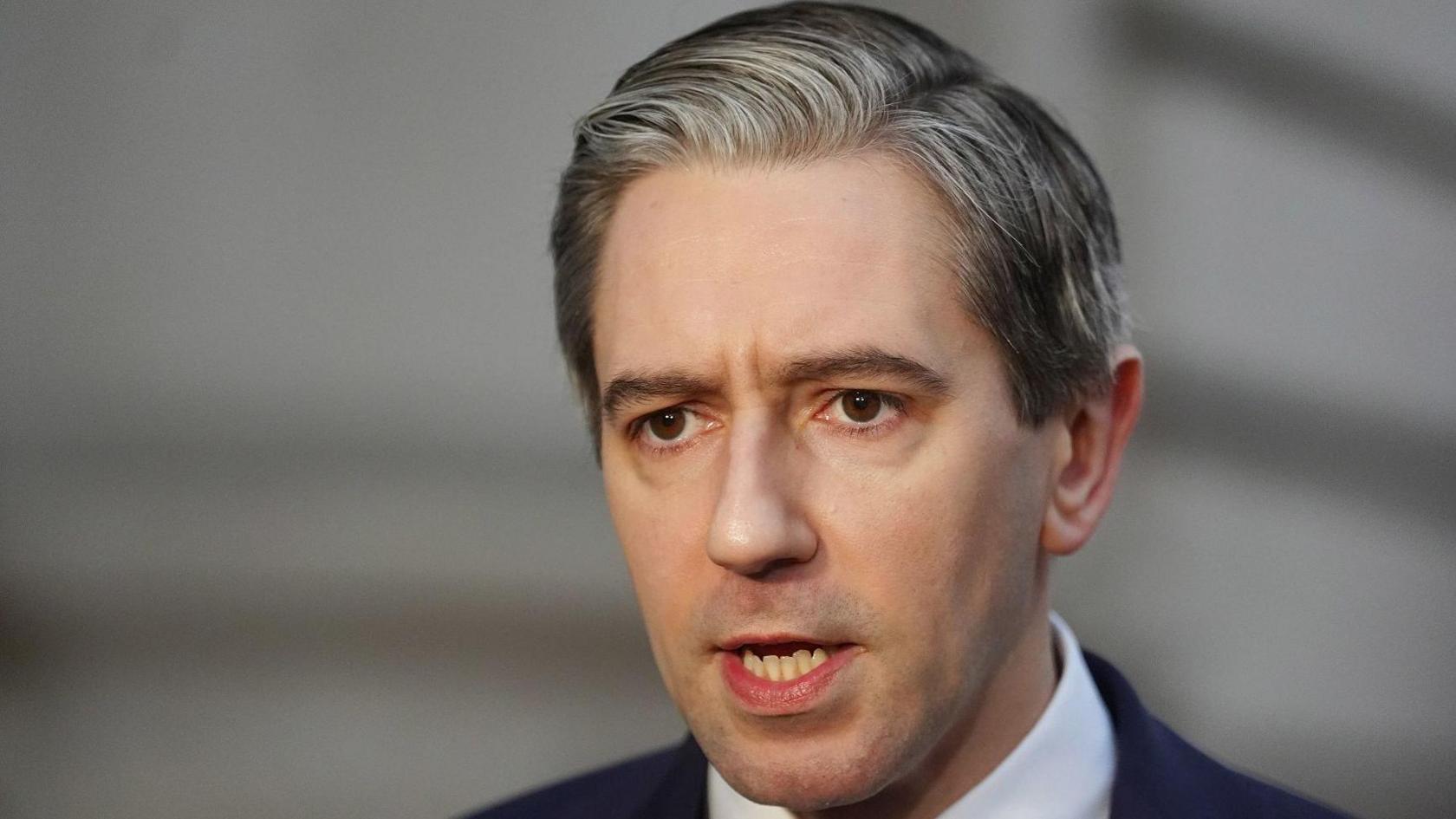 Simon Harris has salt and pepper hair in a side parting and is wearing a dark suit. He has brown eyes and has his mouth slight open in the picture