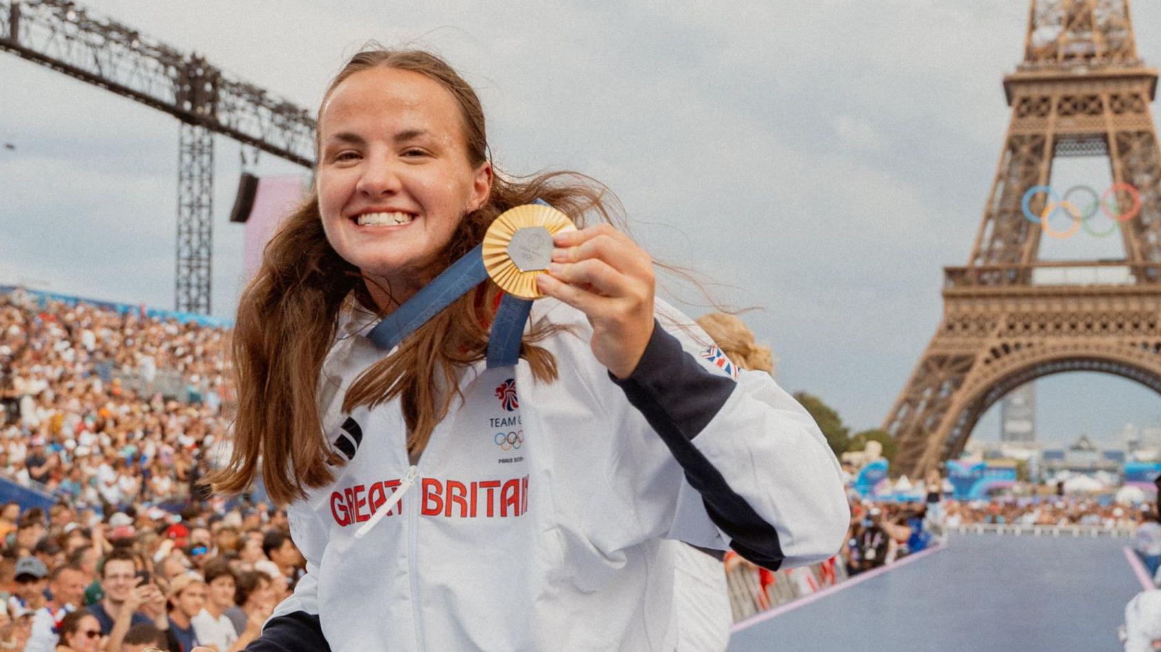 Lauren Henry showing her gold medal to the camera, wearing a white Team GB jacket with part of the Eiffel Tower in the background, which has the five Olympic rings on the front of the landmark at the Paris 2024 games.