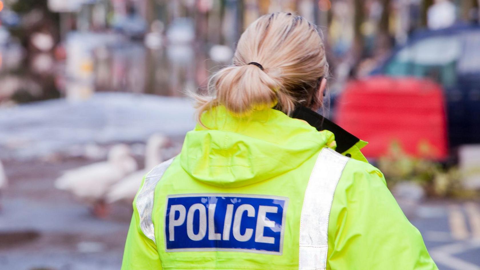 The back of a female police officer. She has blonde hair tied back and is wearing a fluorescent yellow jacket which reads 'police' on the back. The background is fuzzy, but looks to be a street.