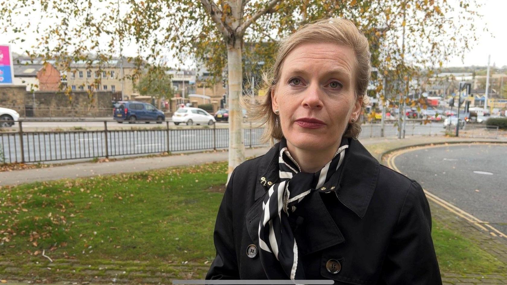 Prof Rosie McEachan, a blonde lady wearing a black coat and a black and white scarf. She is standing against the backdrop of a grass verge and a busy road.