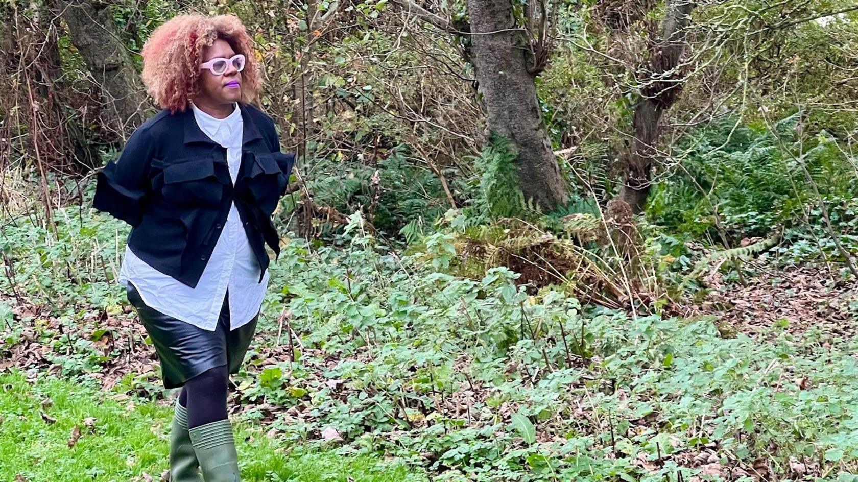 Errollyn Wallen walks through woods in green wellies. She is wearing a black skirt, black tights, a light blue shirt and a dark blue jacket. 