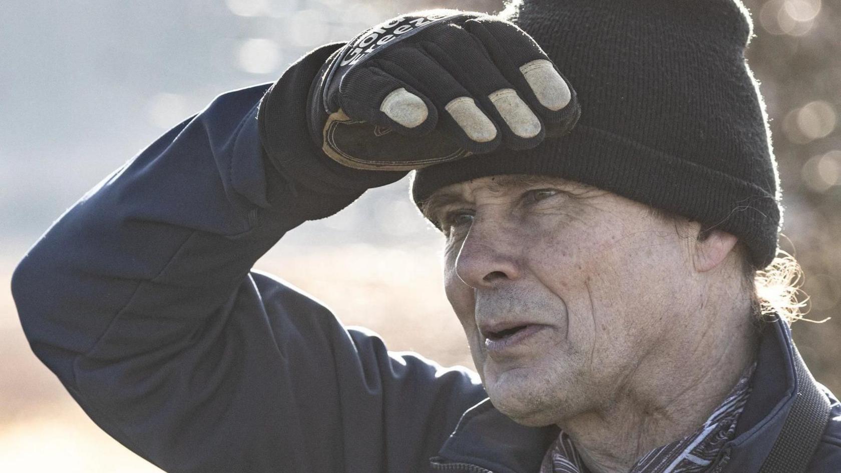 Close up of Dr Vince Lea wearing a black beanie hat, black gloves and a jacket. He is outside and has one hand against his forehead shielding his eyes from the sun as he looks to away from the camera.
