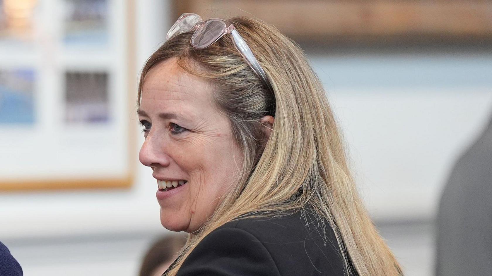 Susan Davy with her back to the camera and looking to her left with a pair of glasses on her head and smiling in a room during a meeting.