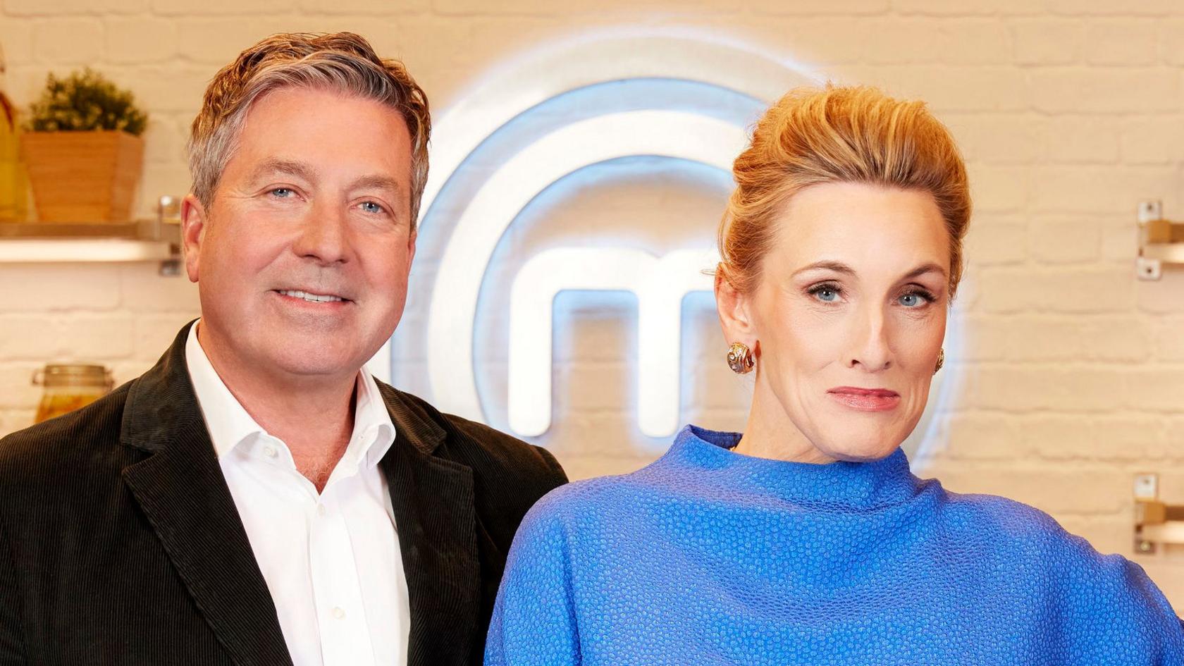 John Torode and Grace Dent pose while looking at the camera in front of the Masterchef logo in a publicity still for Masterchef.