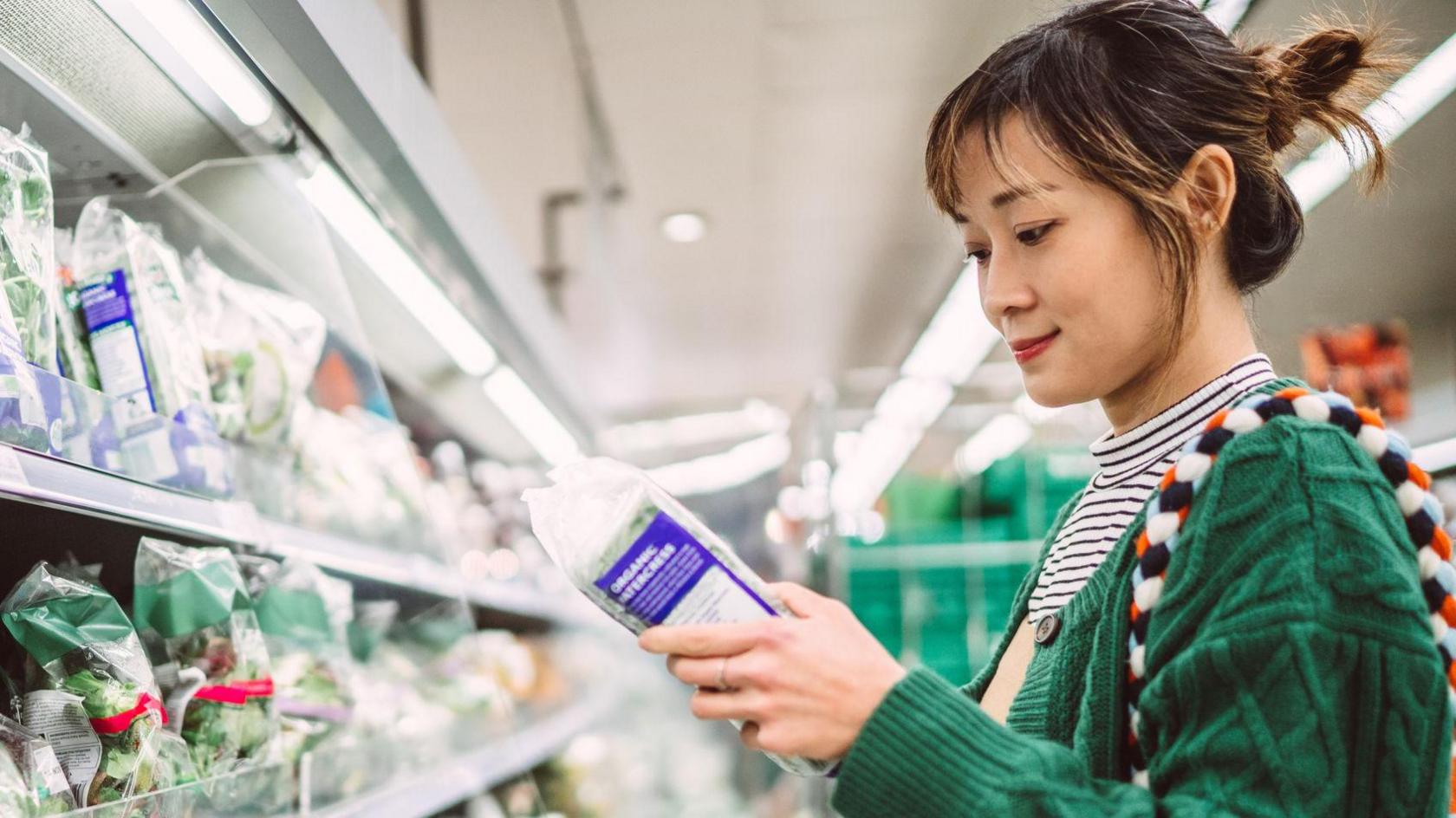 Young woman inspects the packaging of a food item in a supermarket aisle