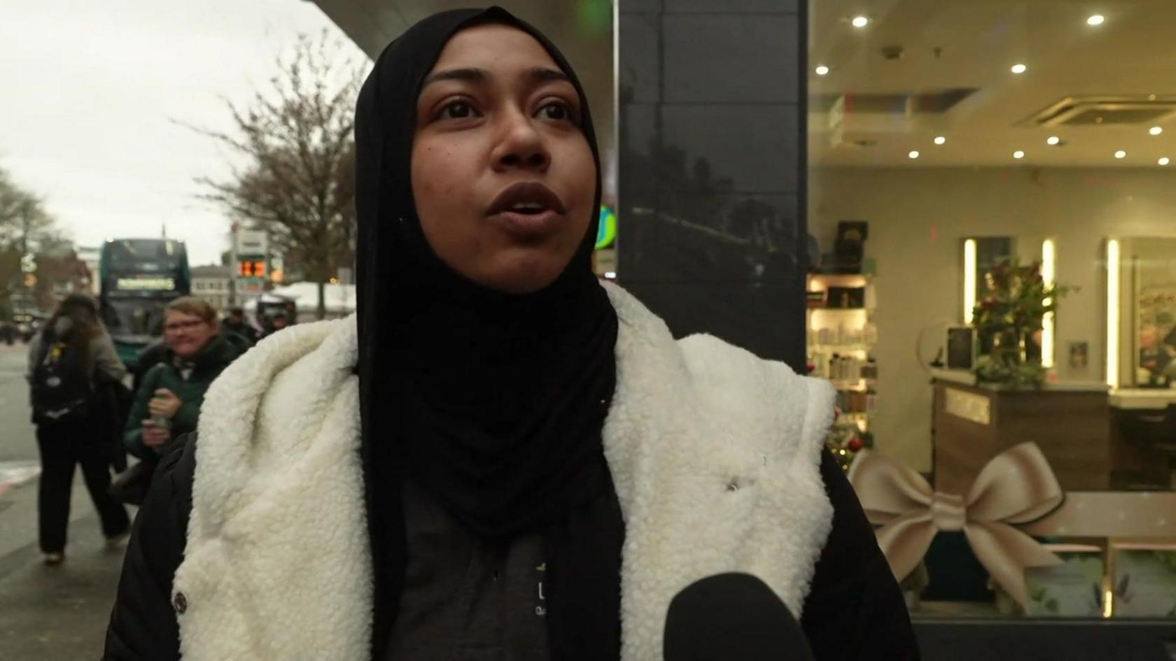 A young woman wearing a dark coat with a furry lining speaks to a BBC reporter, with a black microphone cover in-shot