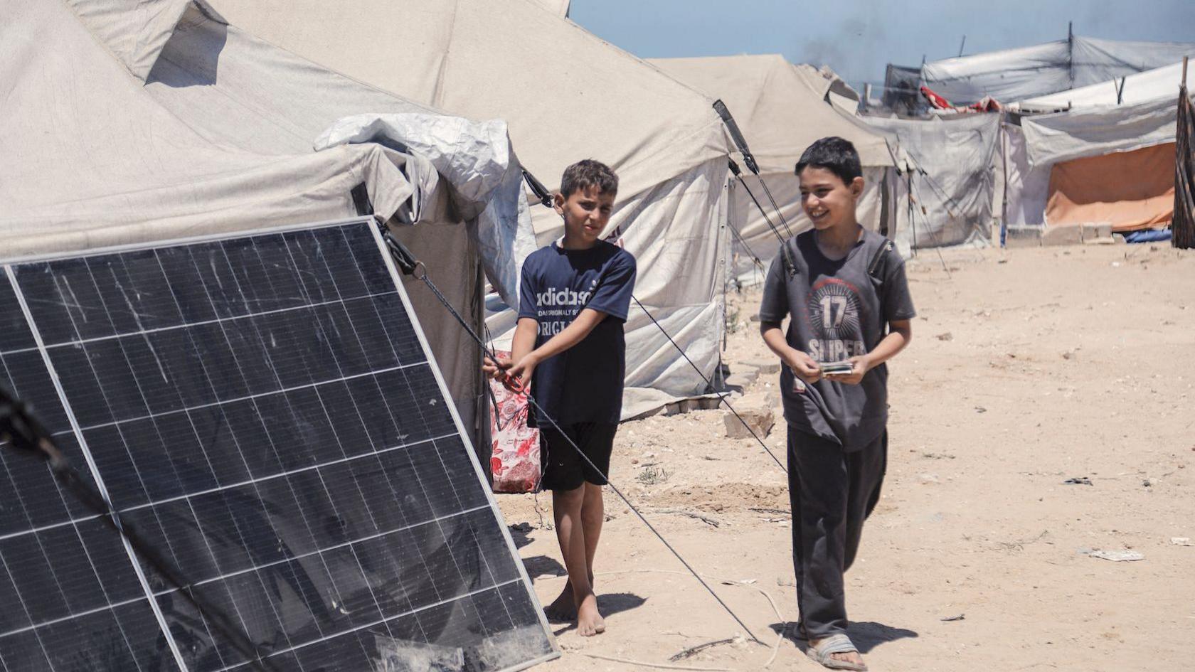 Two boys, one in a navy Adidas T-shirt and shorts and another in a black T-shirt and trousers, stand beside a row of tents, one of which in the foreground has a large solar panel propped up outside it. In the background can be seen some smoke, perhaps from a diesel generator.