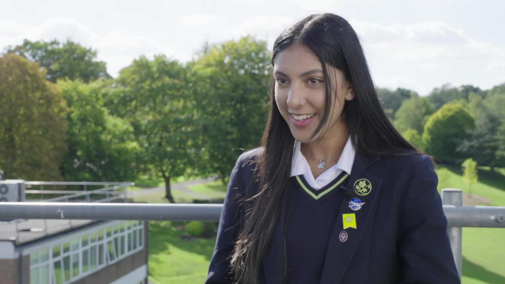 A young girl in school uniform being interviewed on the roof of a school building. Other buildings and green lawns and trees are visible in the background. 
