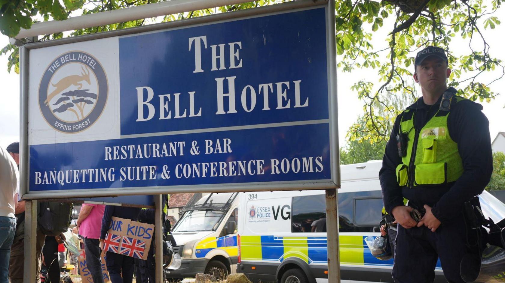 A police officer standing next to a blue sign that says "The Bell Hotel". Behind him are police vans and protesters.