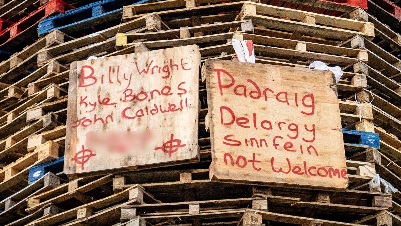 Several names are written on two brown wood placards on a bonfire made up of multi-coloured wooden pallets. 