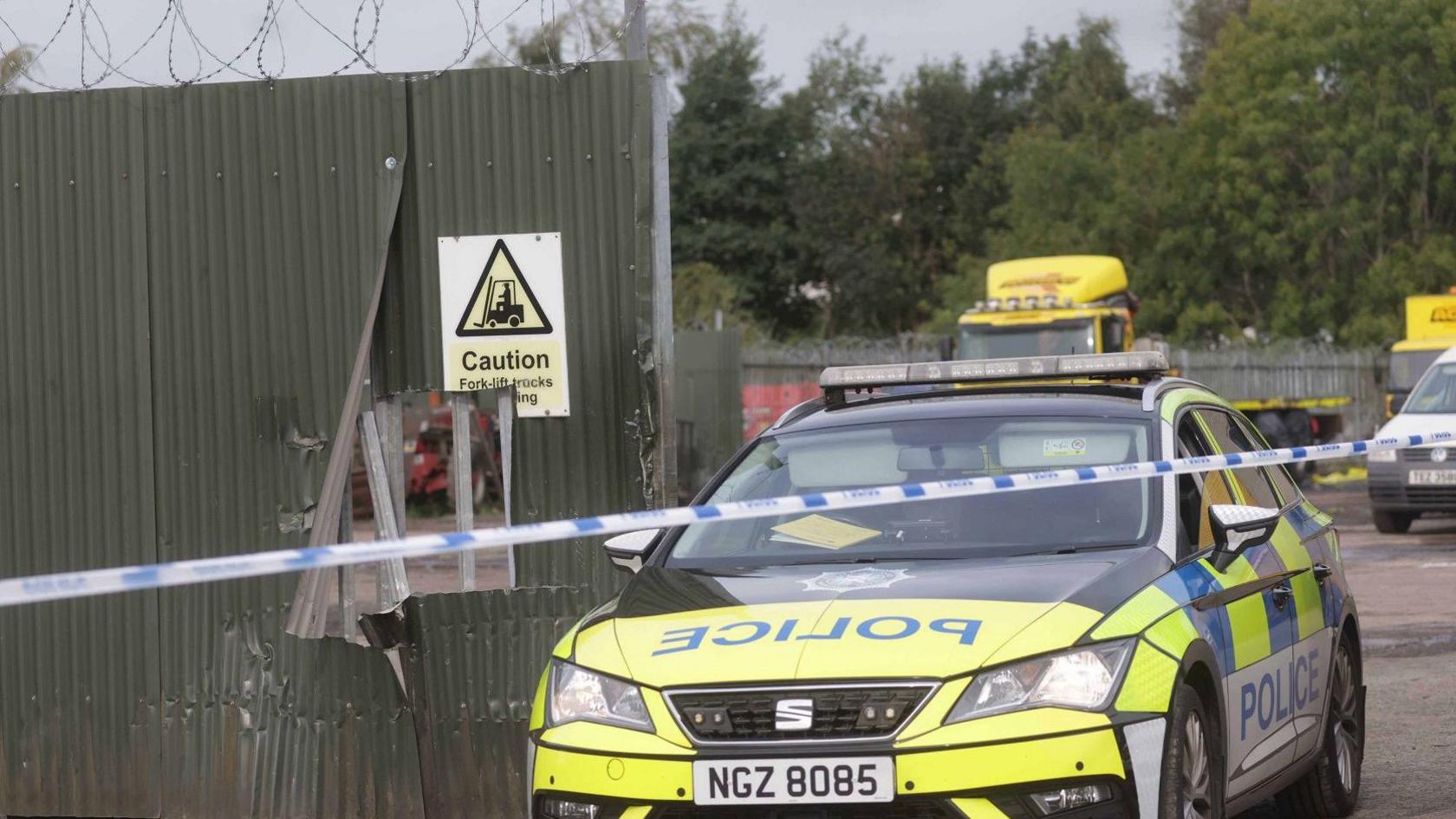 A yellow, blue and white police car is parked behind police tape at an industrial estate. A green fence is beside the car