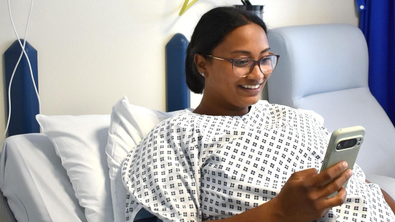 A woman wearing glasses laying on a hospital bed in a gown. She is holding a smart phone in her right hand and reacting happily to what she is looking at one the screen in front of her.