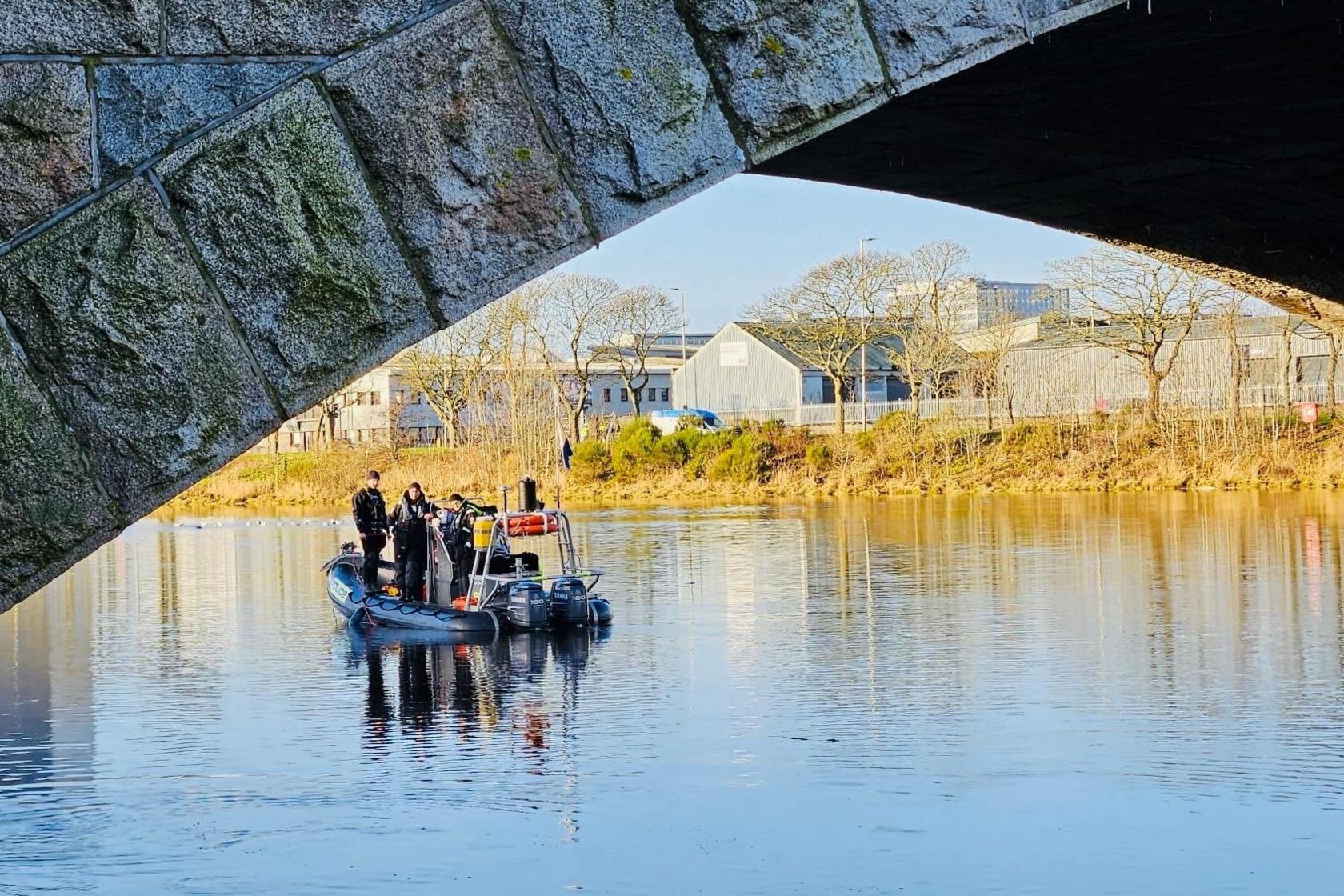 Dinghy with searchers on river, with a bridge arch in the foreground and river banks and trees and buildings in background. 