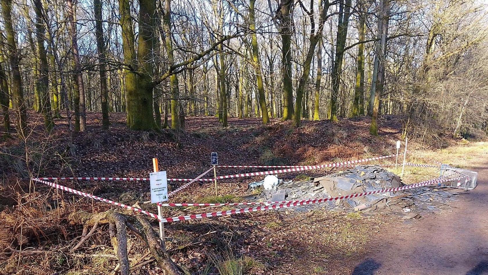 Image shows woodland near Cinderford with an area to the side of the wood path cornered off with red and white tape. In the cornered off area, fly-tipping consisting on brown and black material can be seen in a mound. In the foreground of the image, two shadows can be seen as the picture is taken. 