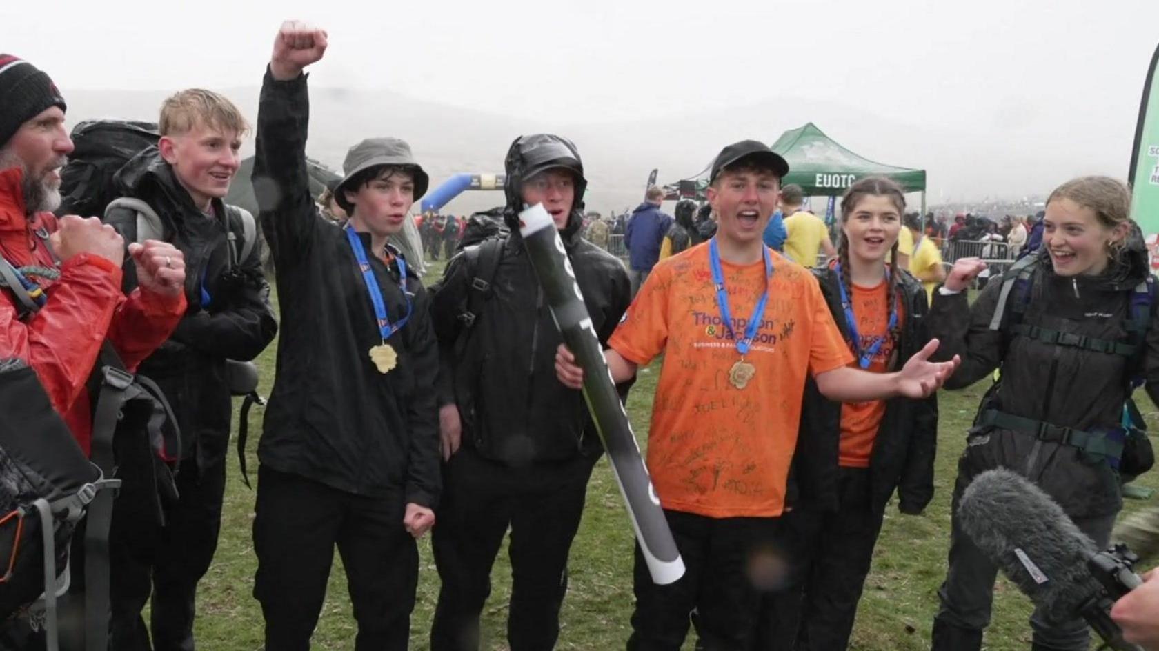 Four young men, two girls and an adult are cheering. The walkers are wearing medals. It is grey and rainy.