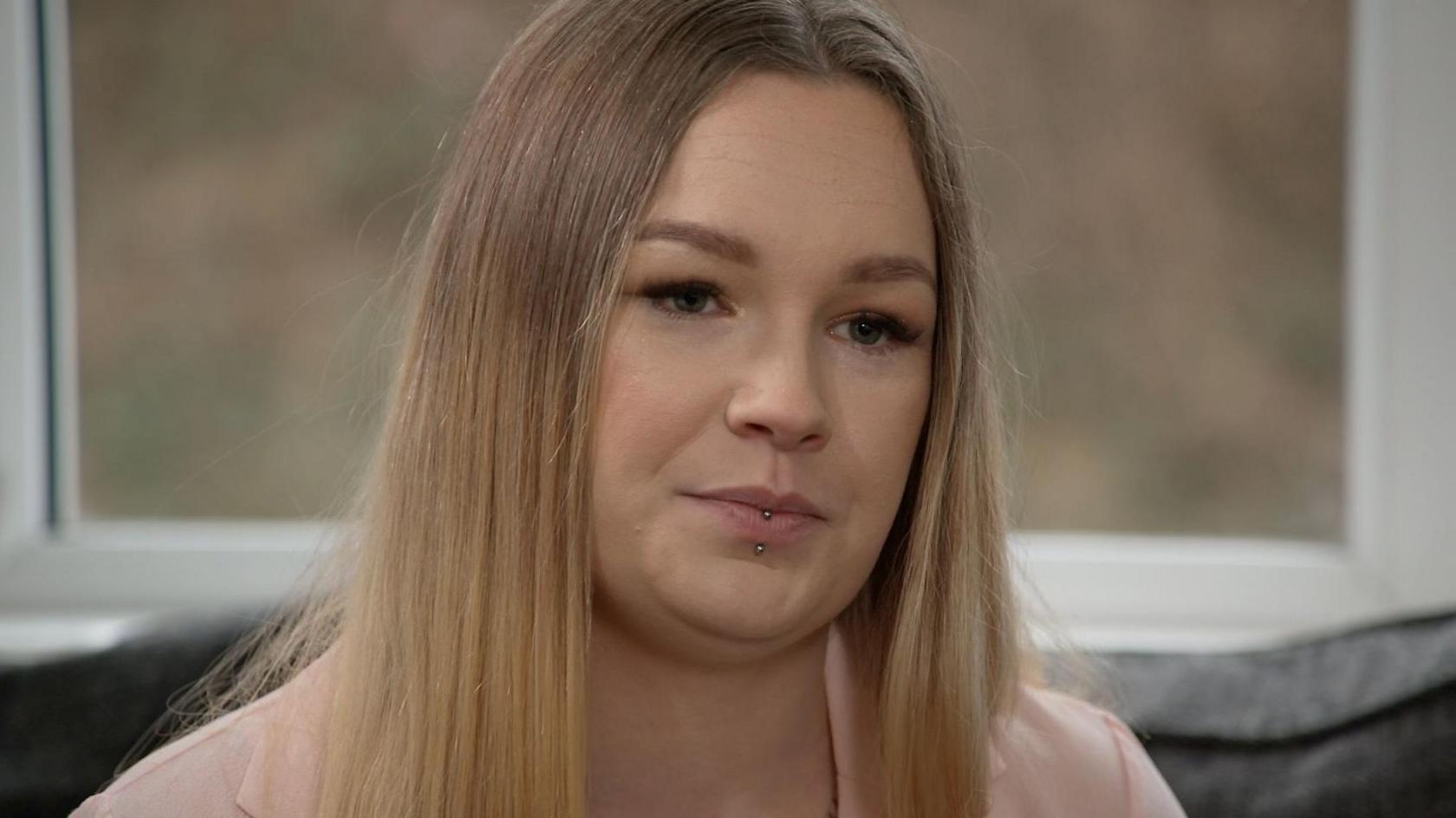 Tia is looking to the right side of the frame, her long straight blonde hair loose around her face. She has a bottom lip piercing and is wearing a pale pink blazer over a white top. The background - the top of a grey sofa and a window - is blurred behind her.