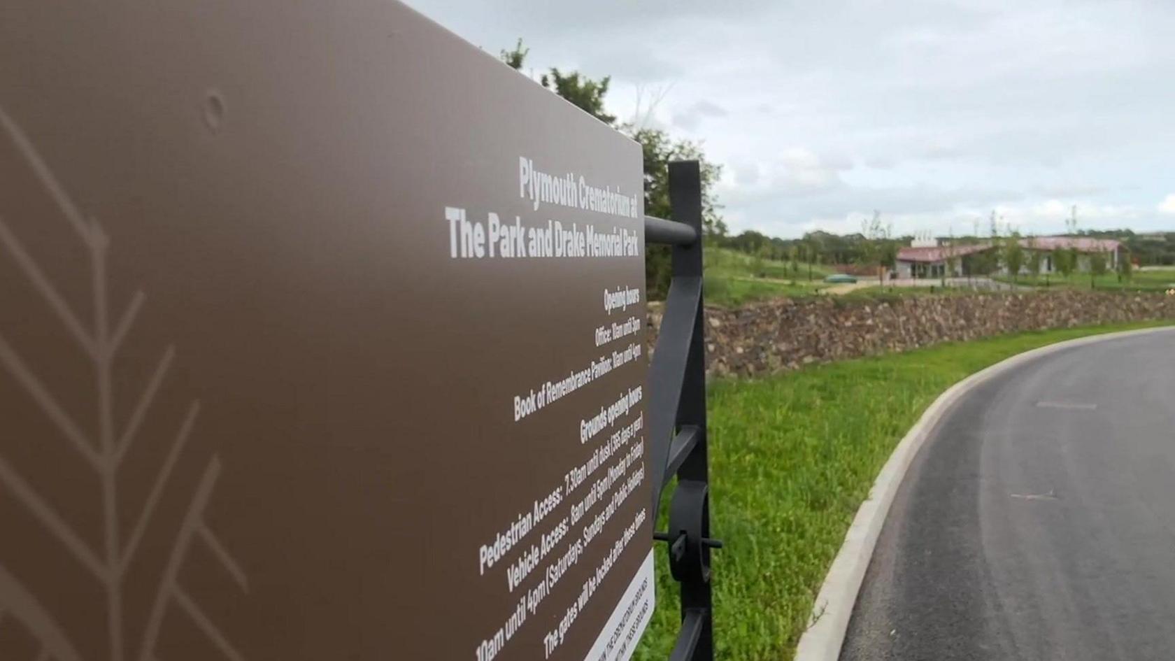 A picture of the entrance to the crematorium. The gate has a brown sign containing white writing. The crematorium can be seen in the background surrounded by grass.