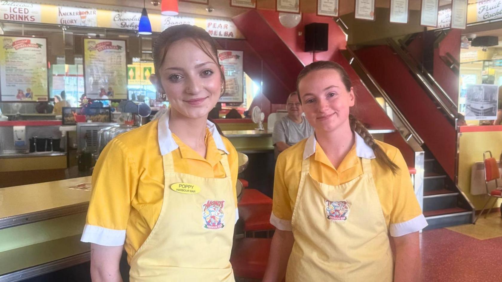Two young women stand in front of the bar at a vintage-style ice cream parlour. They wear yellow 1950s-style uniform.
