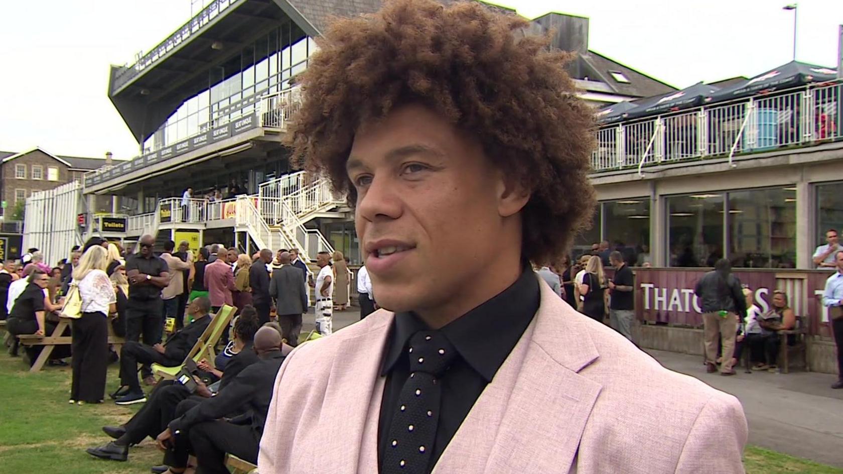Buster Lawrence in a pink suit with a black shirt and polka dot tie standing outside the cricket club with many people behind him.