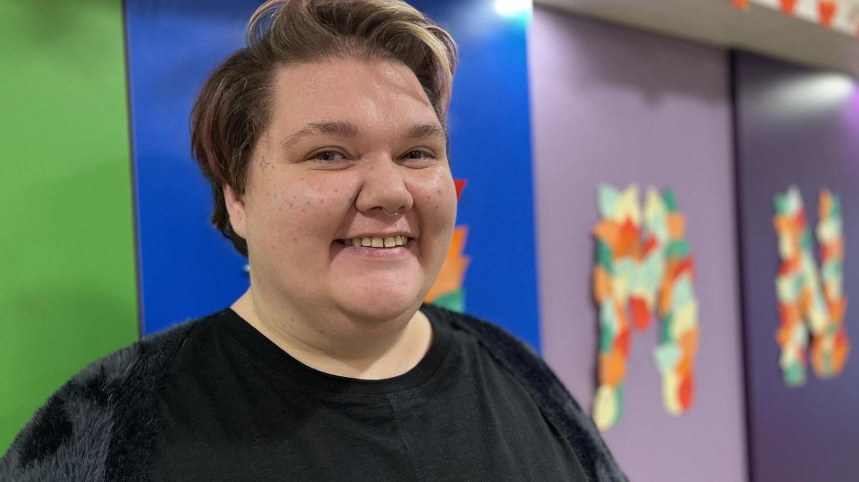 A person with short hair - coloured light brown with flecks of pink - and a small silver nose ring smiles at the camera. They are wearing a black top. The background is colourful with purple, blue and green blocks and large hand-made letter murals in autumnal colours.