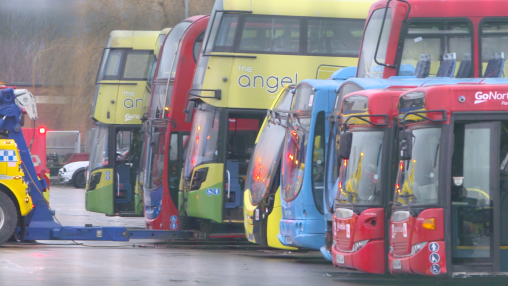 A recovery truck lifts a yellow and green double decker bus off the ground, It is surrounded in the middle of a line of brightly coloured buses.