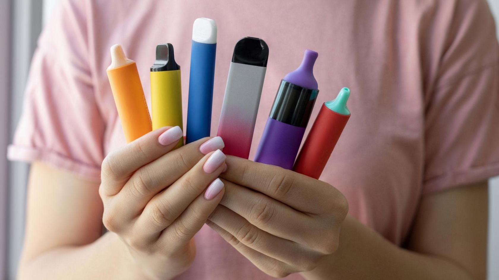 A woman wearing a pink shirt and pink nail polish holding up six colourful, disposable vapes in both her hands. 